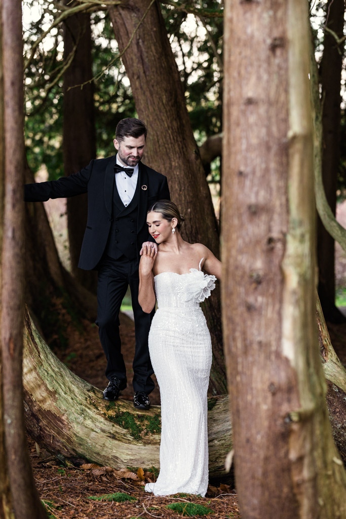 A bride in a white dress and a groom in a black tuxedo pose together among tall trees in a forest on the grounds of Markree Castle