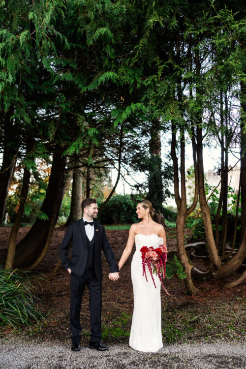 A couple in formal attire holds hands and smiles beneath tall green trees, capturing a romantic moment during their Markree Castle wedding.