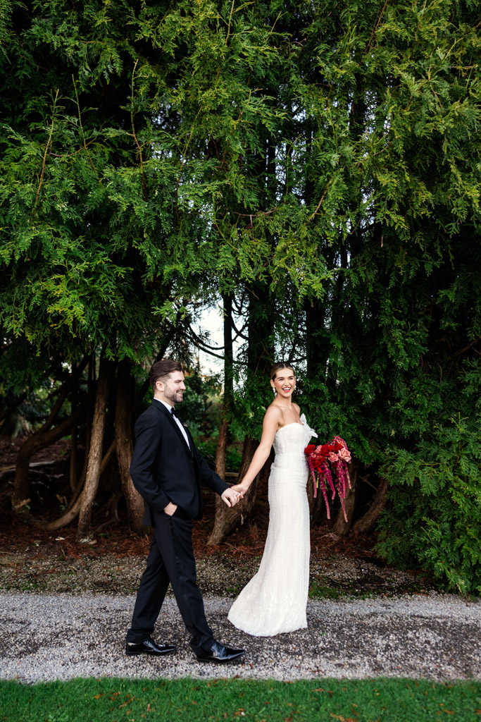 Bride in a white dress and groom in a black suit holding hands, walking outside by green trees during their Markree Castle wedding.