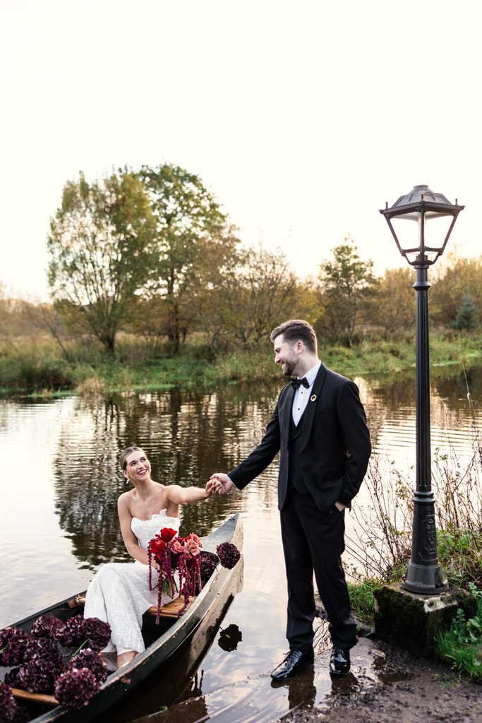 A bride in a boat holds hands with a groom standing by a lake near a lamp post outside Markree Castle
