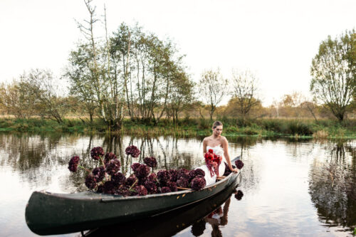A woman in a boat filled with red flowers floats on a calm river surrounded by trees, capturing the fairytale romance of a Markree Castle wedding.