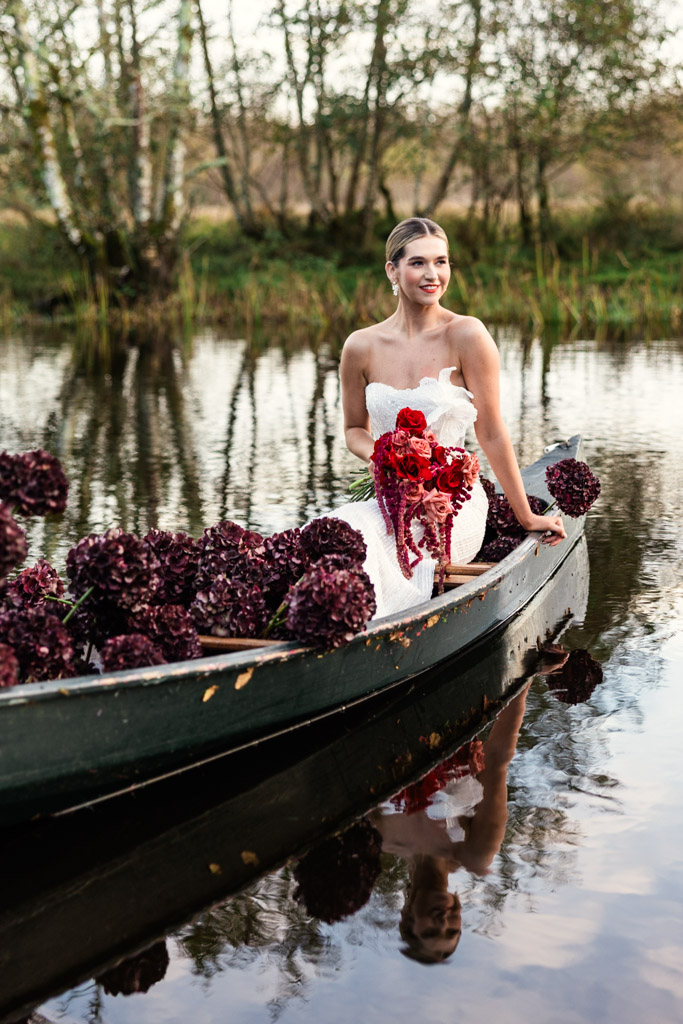 A bride in a white dress holds a red bouquet while sitting in a boat decorated with dark purple flowers during her romantic Markree Castle wedding.