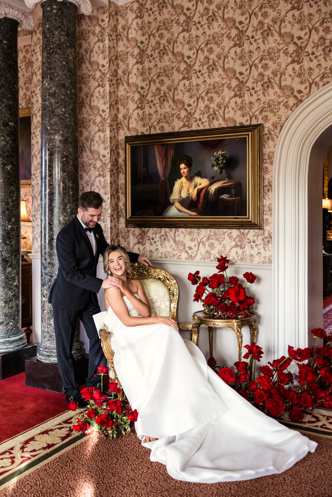 A bride in a white gown sits smiling on a chair, with a groom standing behind her amid red roses indoors at their enchanting Markree Castle wedding.