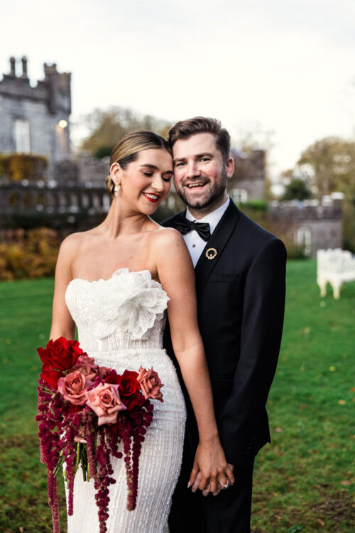 A bride and groom stand smiling outdoors at their Markree Castle wedding; she holds a bouquet of red roses and wears a strapless white dress.