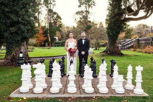 A bride and groom stand behind a large outdoor chess set on the grassy lawn of a garden, celebrating their Markree Castle wedding.