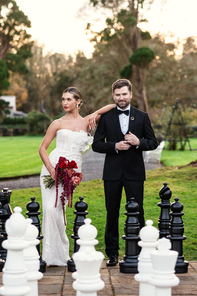 Bride in a white dress and groom in a tuxedo stand as king and queen of a giant chess set outdoors at Markree Castle wedding