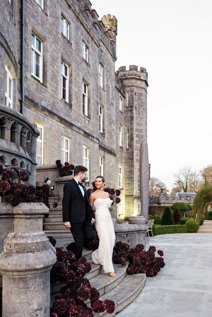 Bride and groom in formal attire walk down stone steps outside Markree Castle, surrounded by dark red flowers
