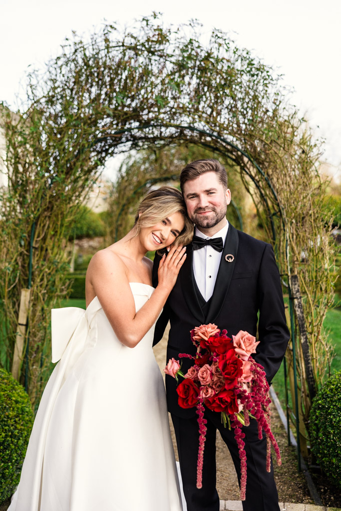 Portrait of bride in a white gown and groom in a black tuxedo holding red flowers, standing under a garden arch at Markree Castle