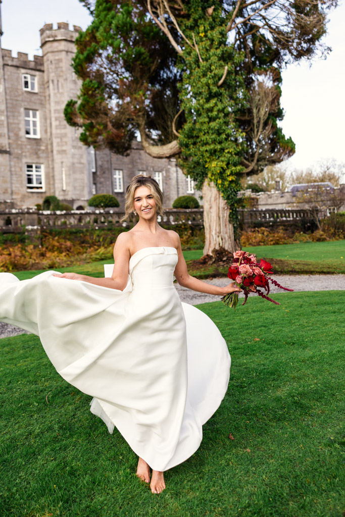 Barefoot bride in a white dress holding a bouquet, smiling outside Markree Castle with a lush green lawn and trees—a dreamy Markree Castle wedding moment.