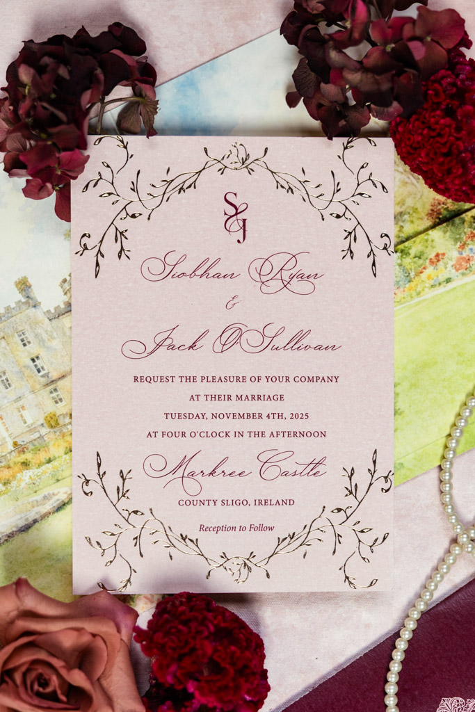 Rose-pink wedding invitation with floral border, surrounded by red flowers, a pearl necklace, and a watercolor painting for Markree Castle wedding