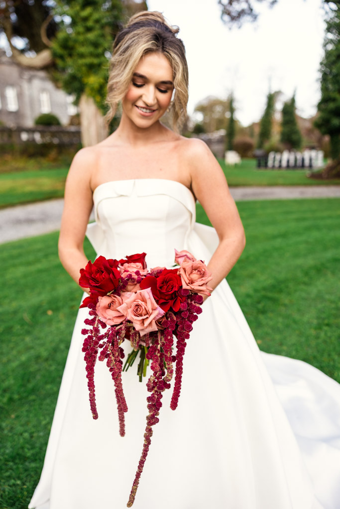 A bride in a white dress holds a bouquet of red and pink flowers, standing on green grass outside Markree Castle