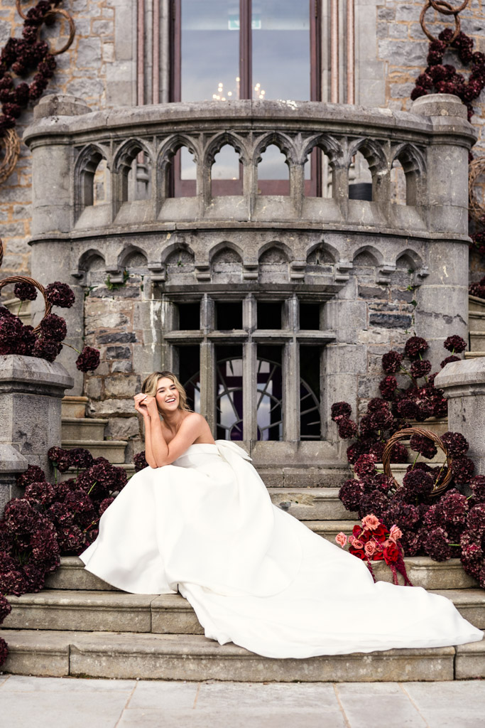 A smiling bride in a white gown sits on stone steps, surrounded by dark red flowers outside Markree Castle