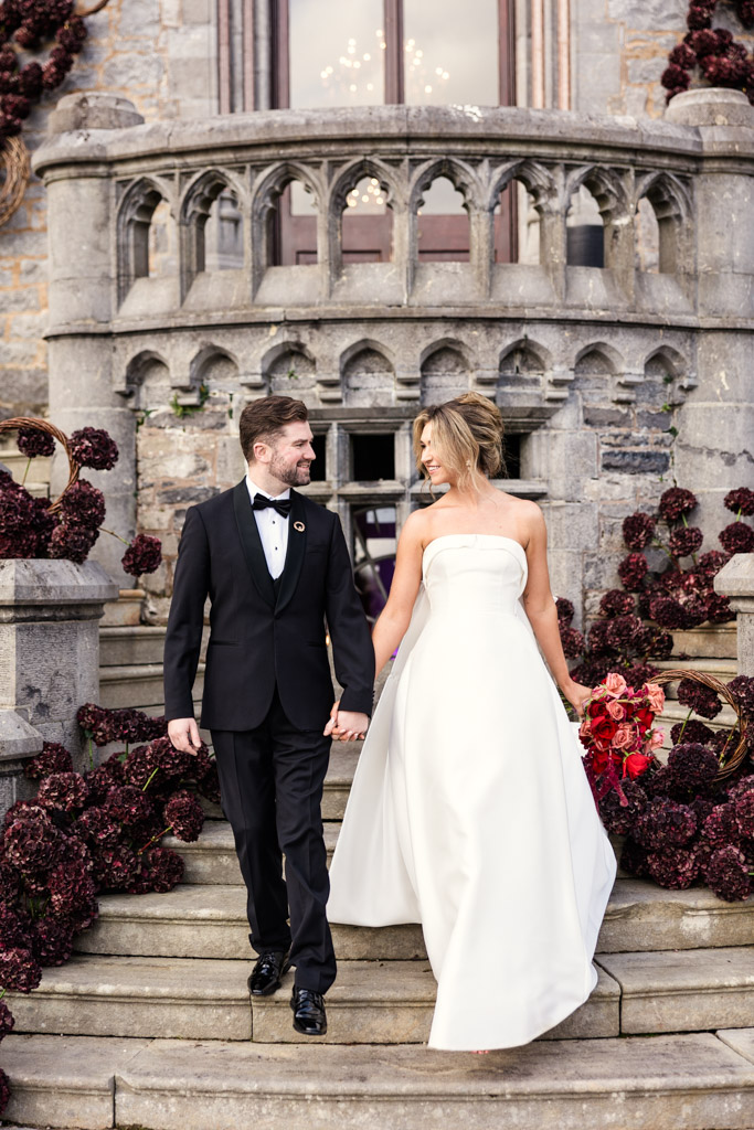 A bride and groom in formal attire hold hands and smile while descending stone steps adorned with dark flowers at Markree Castle
