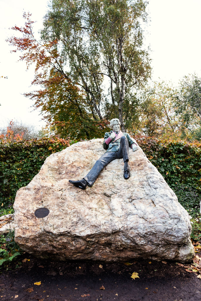 Oscar Wilde Monument in Dublin depicting WIlde reclining on a large rock, surrounded by autumn trees and greenery