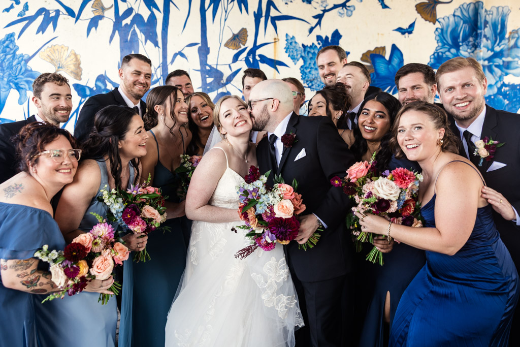 A bride and groom kiss, surrounded by a joyful wedding party with floral bouquets, in front of a blue floral mural at Ping Tom Memorial Park