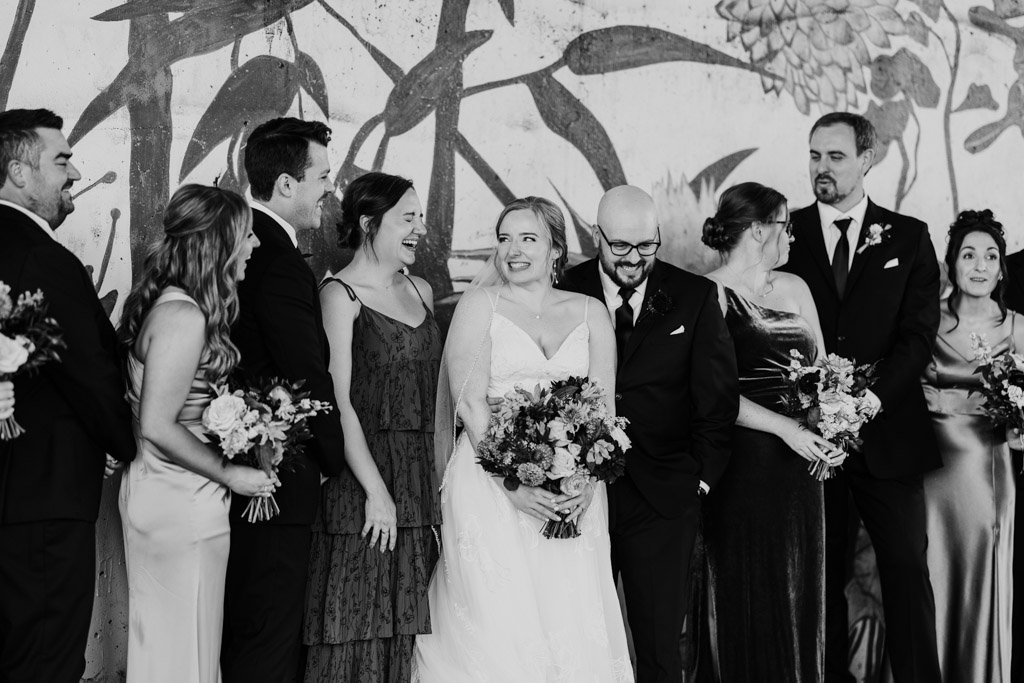 Candid black and white photo of wedding party smiling and laughing together in front of a floral mural at Ping Tom Memorial Park