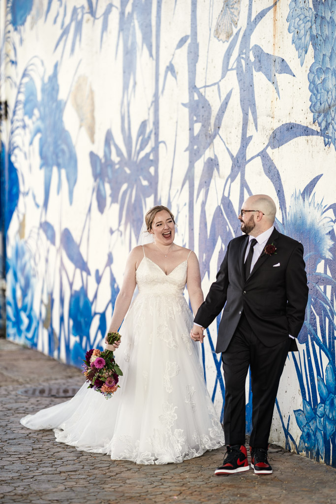 Bride and groom holding hands, smiling, walking in front of a blue and white floral mural at Ping Tom Memorial Park