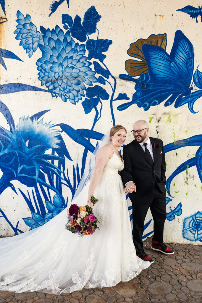 Bride and groom smiling in front of a blue floral mural at Ping Tom Memorial Park; the bride holds bouquet and her veil and train flows behind