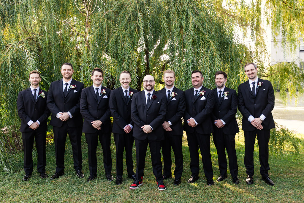 Portrait of groom and groomsmen pose outdoors in front of green trees at Ping Tom Memorial Park, the groom wearing red sneakers