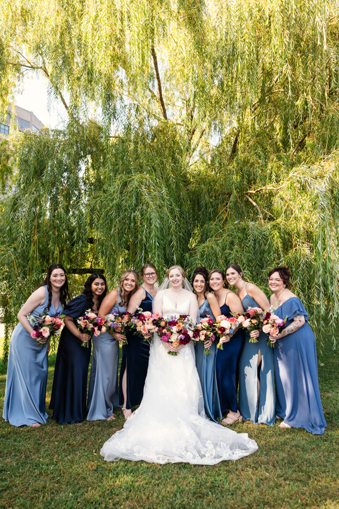 Portrait of bride in white with bridesmaids in blue dresses, holding colorful bouquets, beneath a weeping willow at Ping Tom Memorial Park