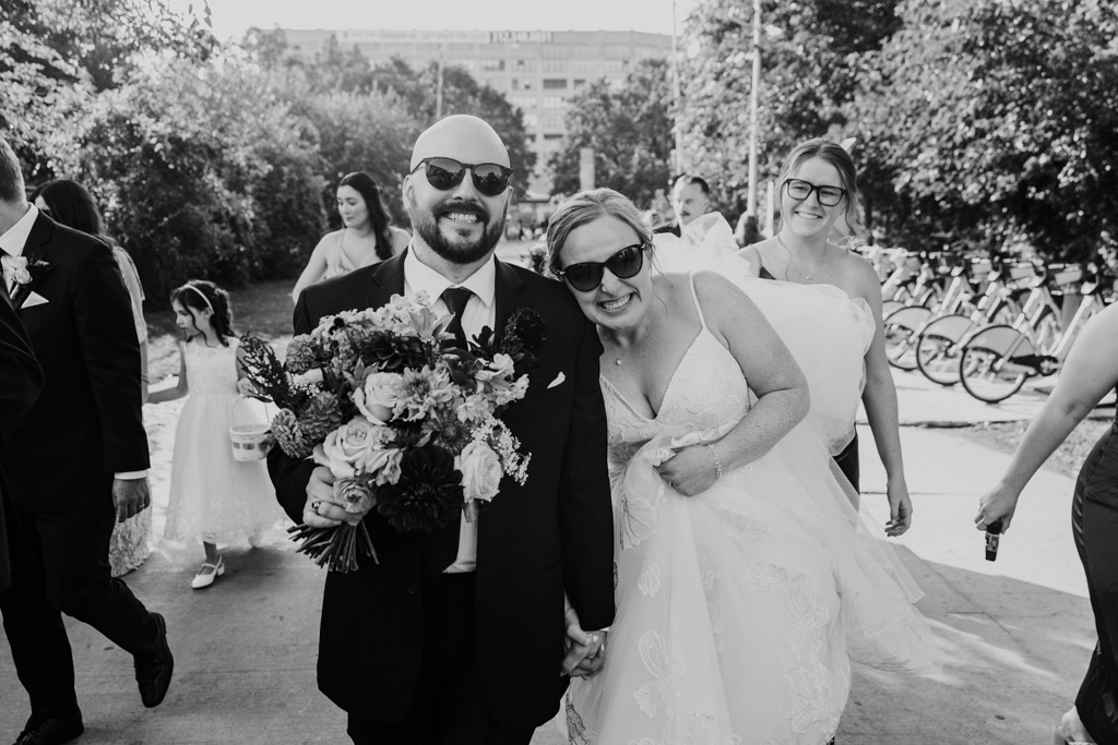 Black and white photo of smiling bride and groom wearing sunglasses while walking outdoors at Ping Tom Memorial Park, holding hands, with friends and family around them