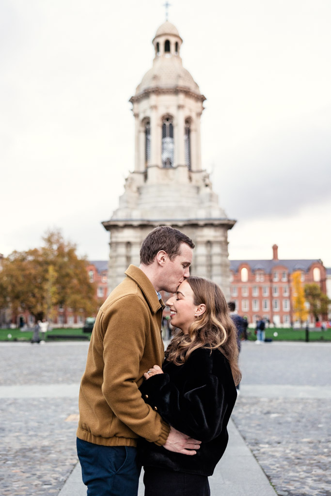 Man kisses his partner’s forehead as they embrace in front of a historic stone tower, on the grounds of Trinity College in Dublin