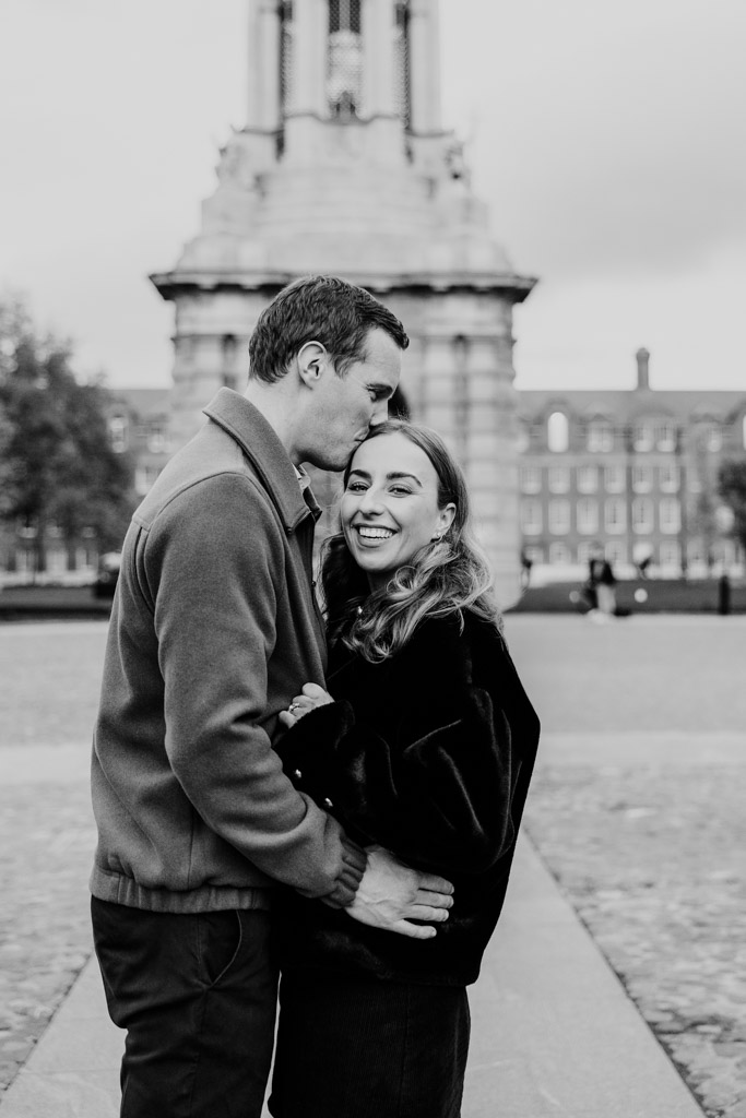 Blacka dn white photo of man kisses his partner’s forehead as they embrace in front of a large stone tower at Trinity College, Dublin