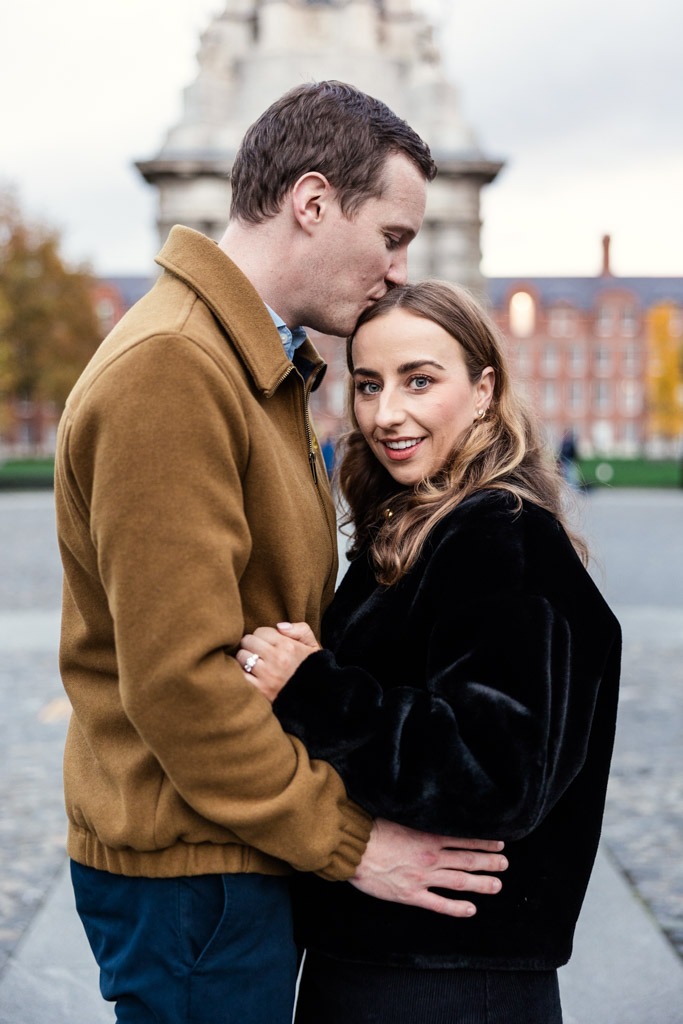 Man kisses his partner's forehead while they stand on the grounds of Trinity College during their Dublin engagement session with school buildings in the background