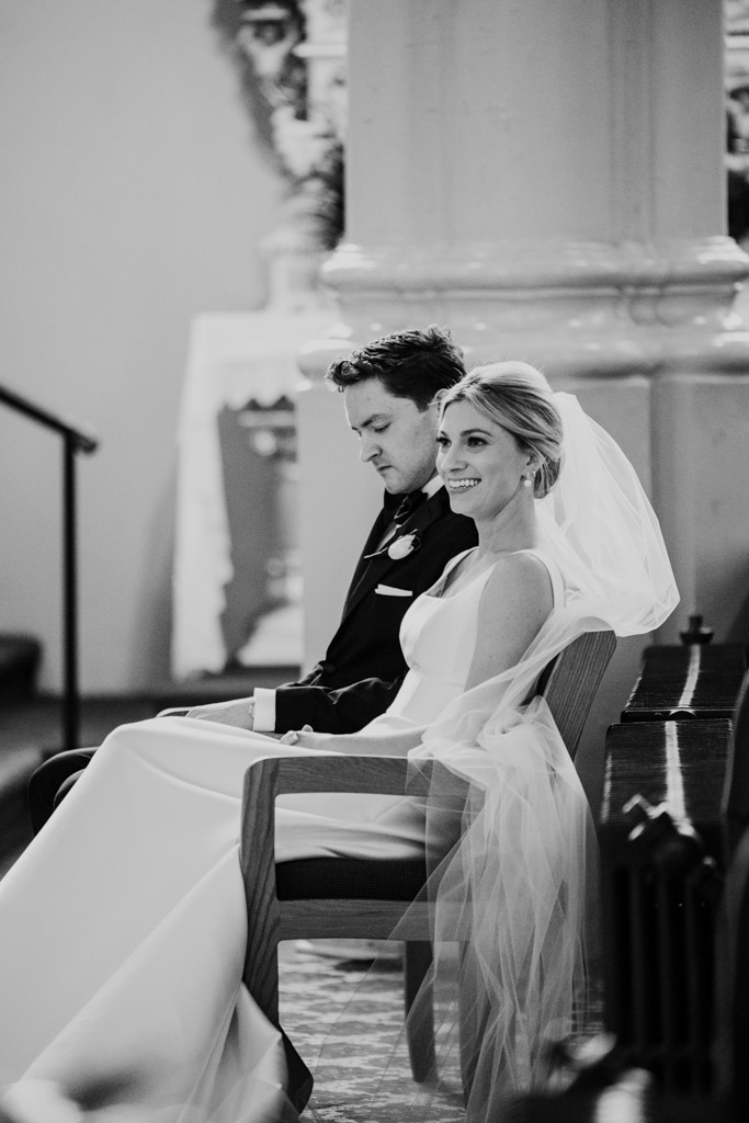 Black and white photo of bride and groom, dressed in wedding attire, sitting side by side in St. Michael's Church as the bride smiles softly
