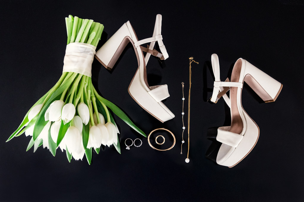 Bride's white high heels, a bouquet of white tulips, rings, bracelets, and a necklace arranged on a black background