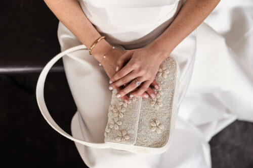Bride in white dress with hands folded on a beaded purse, showing a ring and delicate bracelet