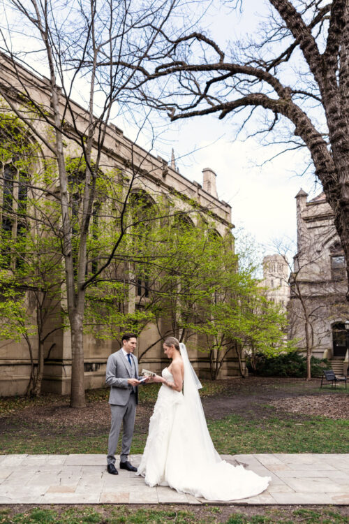 A bride and groom stand facing each other outside, holding hands near a historic stone building and trees.