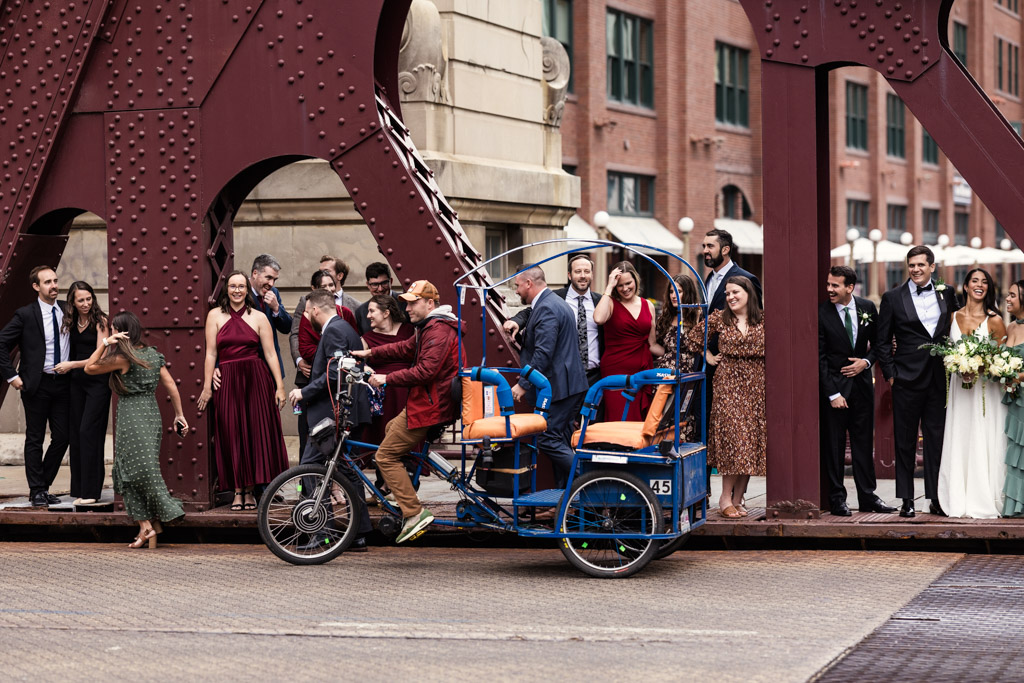 A wedding party poses on a city bridge as a blue pedicab with passengers passes in front of them, capturing the lively spirit of a Bottom Lounge wedding.