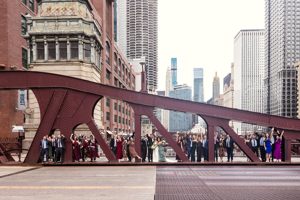 A wedding party poses on a red bridge in a city with tall buildings in the background, capturing the urban charm of a Bottom Lounge wedding.