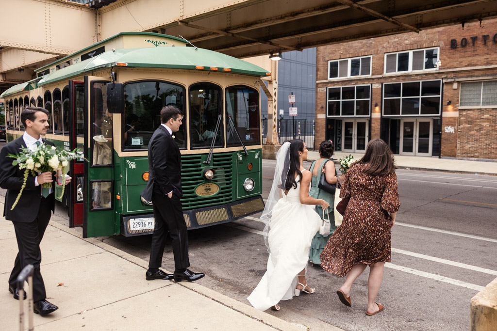 Bride, groom and friends board a vintage green trolley on a Chciago street under an elevated train track, heading to their Bottom Lounge wedding