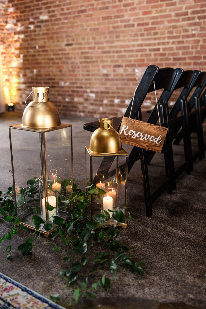 Two gold lanterns with candles and greenery sit beside black chairs—one marked with a 