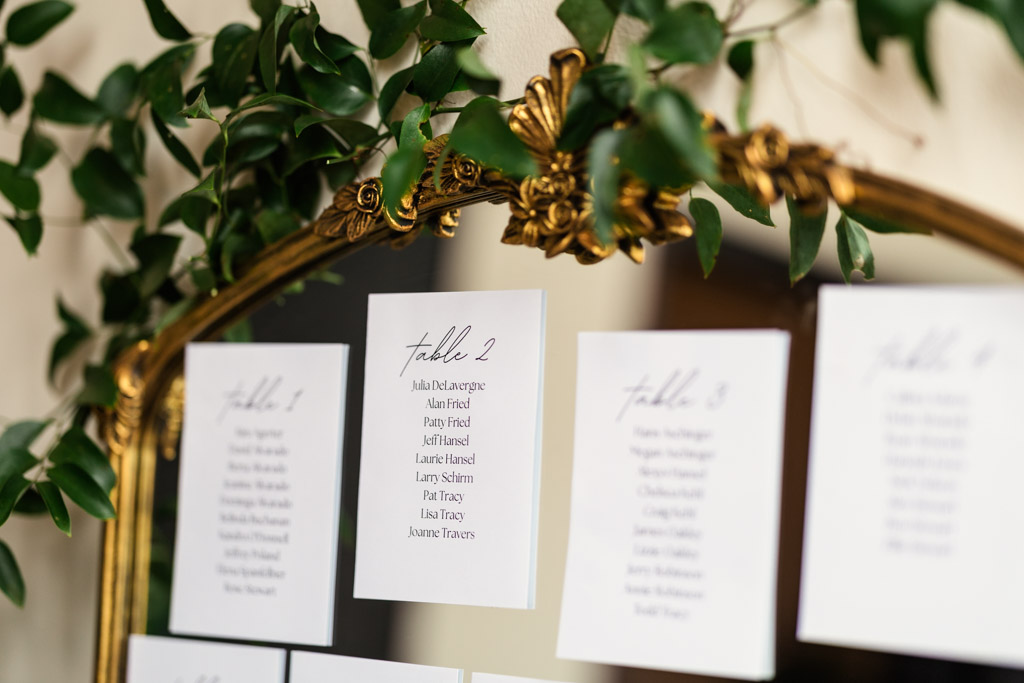 White cards with table assignments are displayed on a gold-edged mirror decorated with green leaves for a Bottom Lounge wedding reception