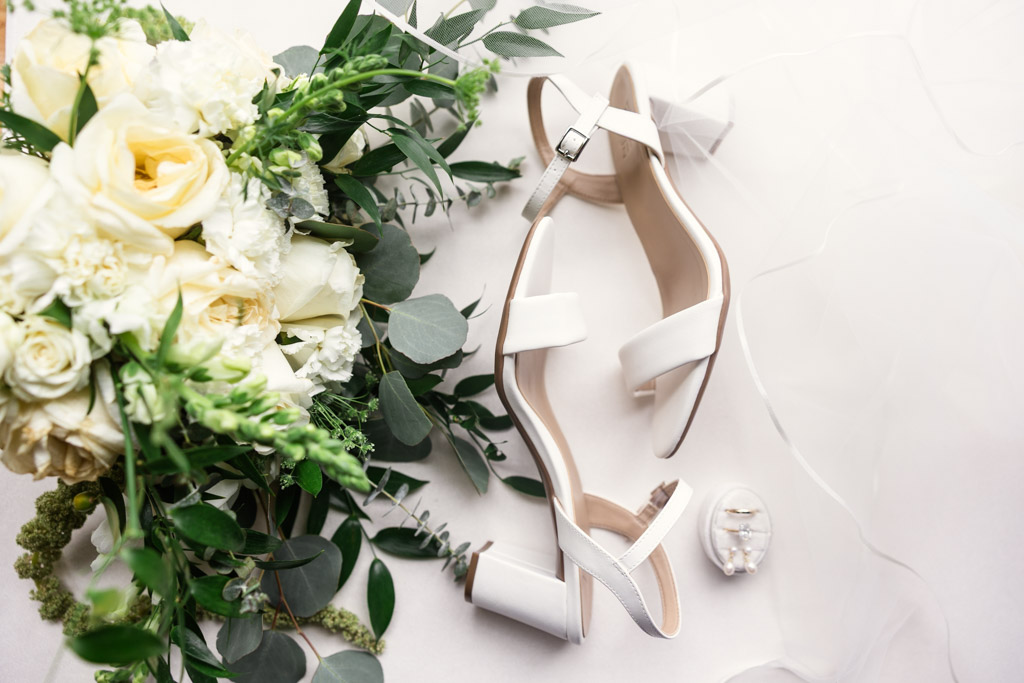 White bride's shoes, a bouquet of white flowers, and a ring box arranged on a light background for Bottom Lounge wedding celebration