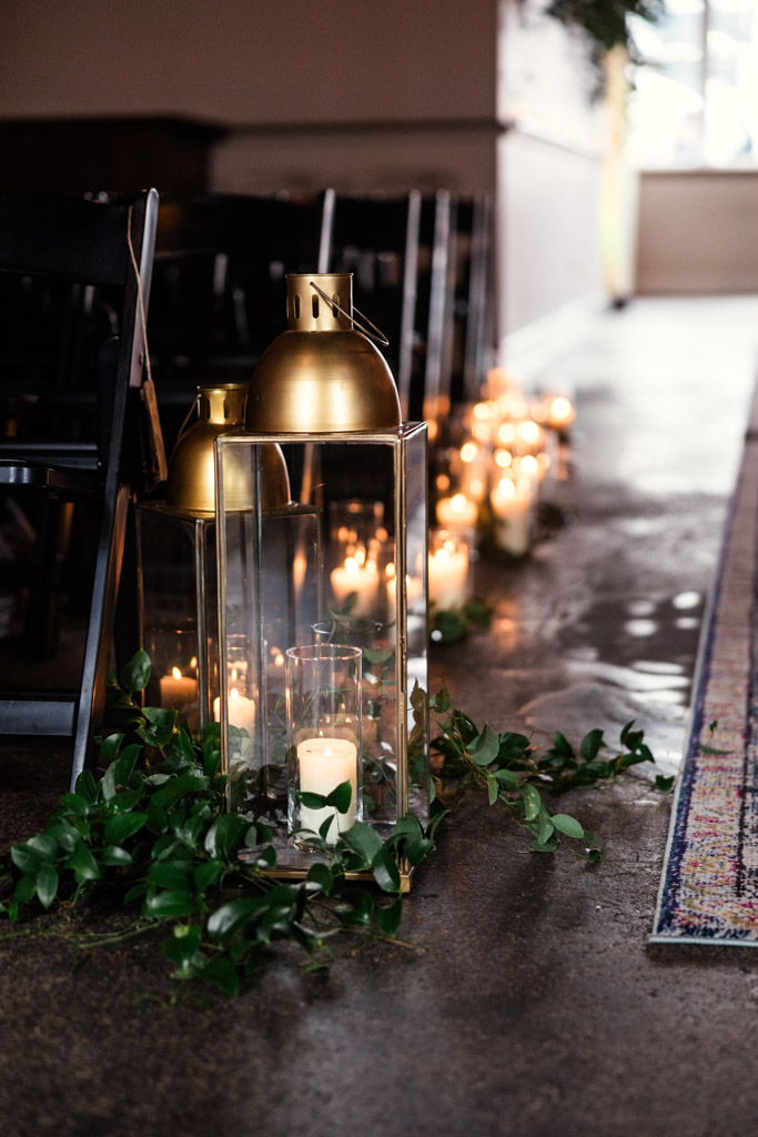 Glass lanterns with candles and greenery line an aisle next to black chairs for a Bottom Lounge wedding