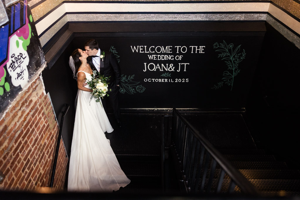 Bride and groom kiss on a staircase beside a sign welcoming guests to their Bottom Lounge wedding