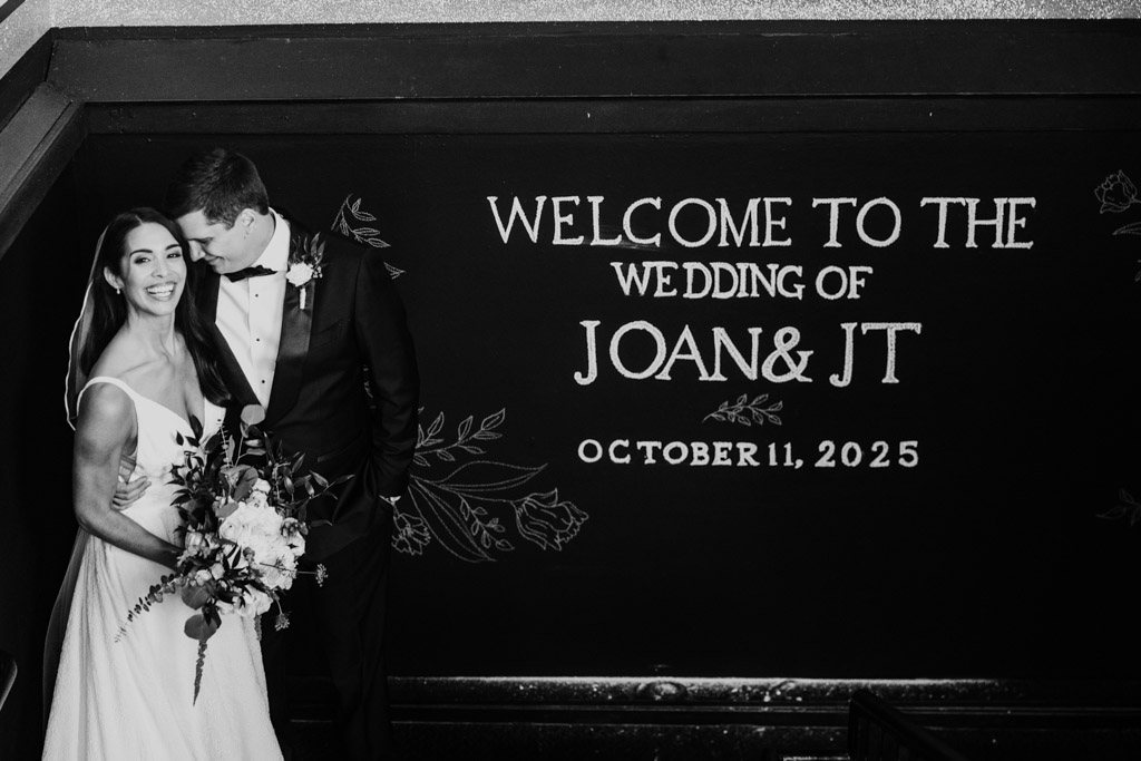 A couple stands smiling on stairs beside welcome sign for their wedding at Bottom Lounge in Chicago