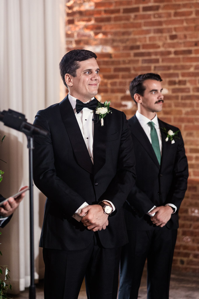 Groom in tuxedo watches bride enter their Bottom Lounge wedding ceremony in front of a brick wall