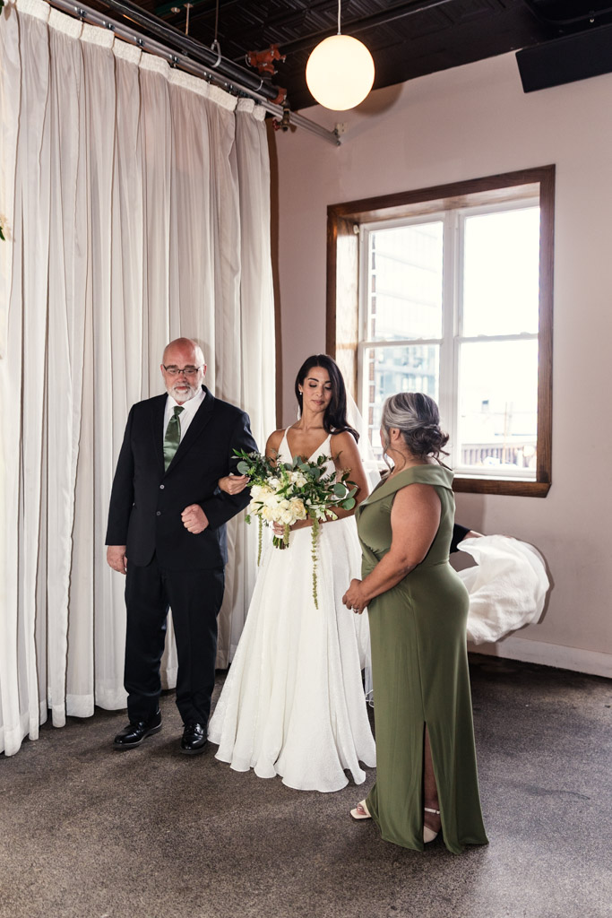 Bride holding bouquet stands with her parents by a window with white curtains at her Bottom Lounge wedding