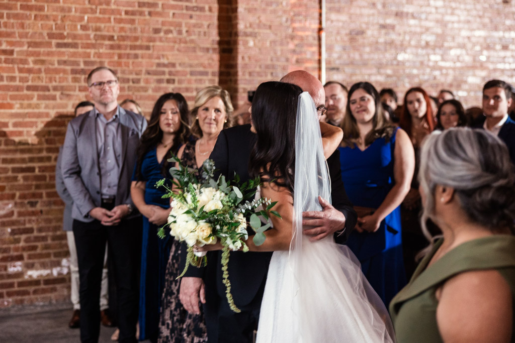 A bride in a white dress hugs her father as guests watch, standing in front of a brick wall at their Bottom Lounge wedding