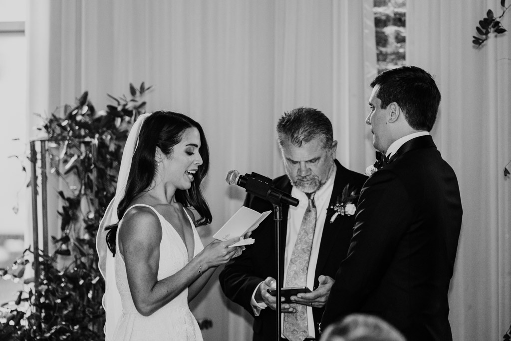 Black and white photo of bride reading vows to groom at their Bottom Lounge wedding ceremony, officiant standing between them, all in formal attire