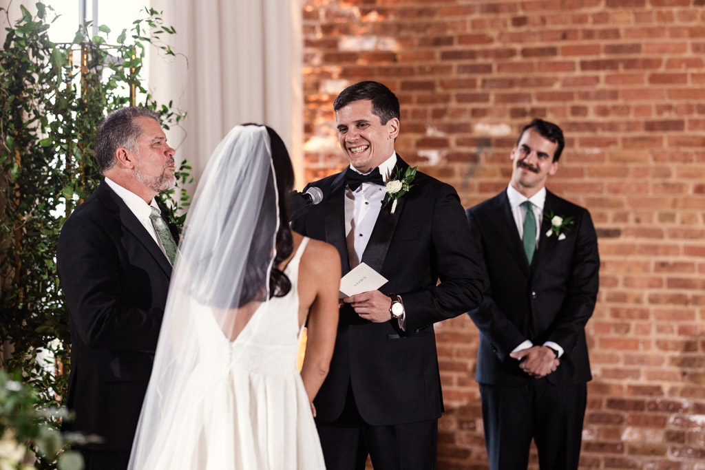 A groom smiles at his bride as he reads his vows during a Bottom Lounge wedding ceremony, with an officiant and groomsman nearby