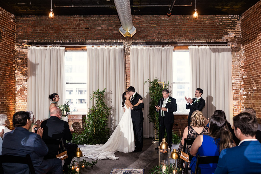 A bride and groom kiss at their indoor Bottom Lounge wedding ceremony as guests watch and clap
