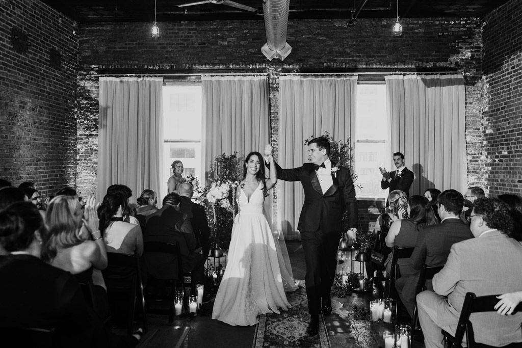 Black and white photo of bride and groom walking down the aisle together, smiling, as guests seated on both sides applaud iafter their Bottom Lounge wedding ceremony