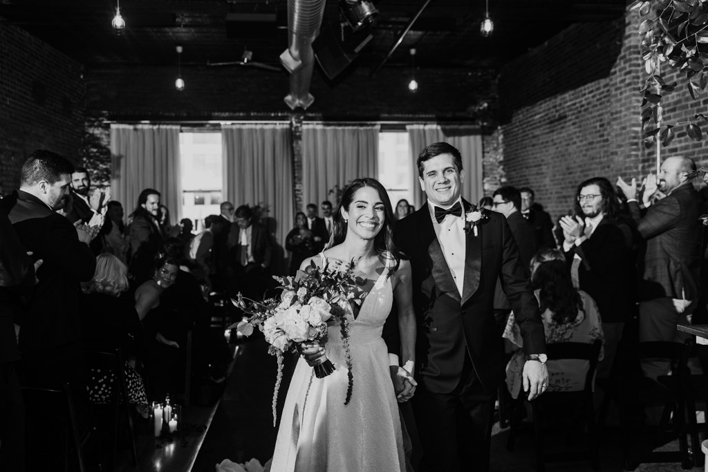 Black and white photo of happy bride and groom walking down the aisle at their Bottom Lounge wedding, surrounded by applauding guests