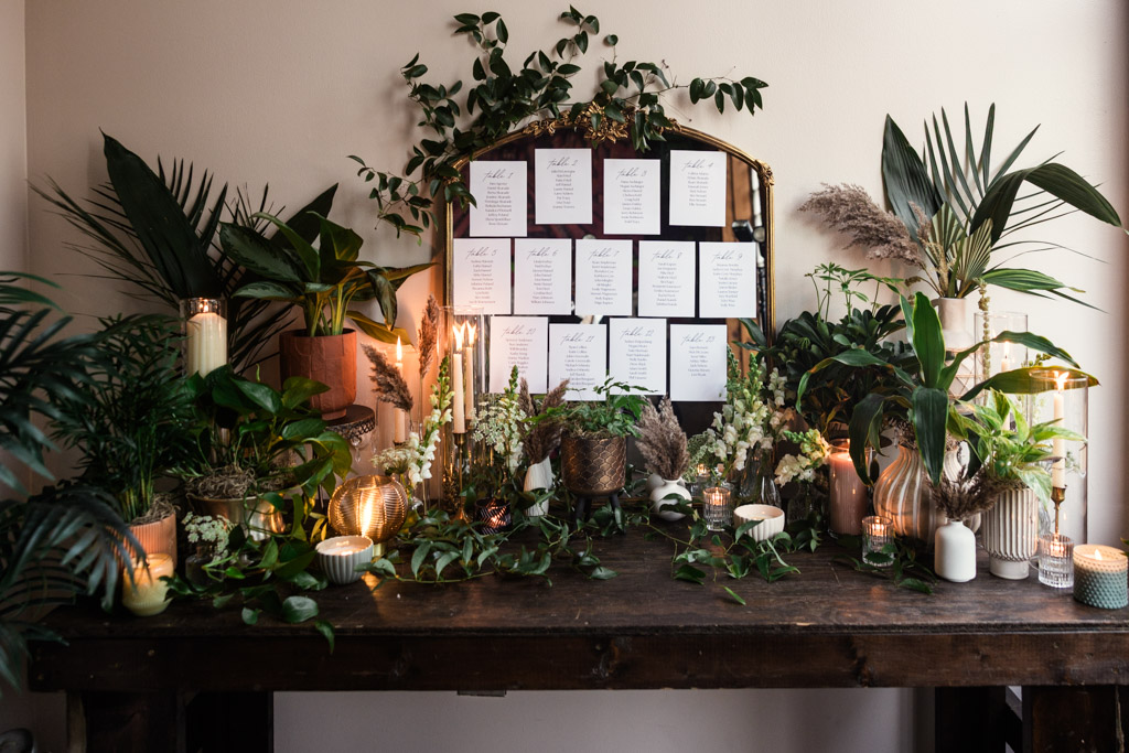A wooden table with candles, green plants, and table assignment cards displayed on a mirror against a light wall for Bottom Lounge wedding reception